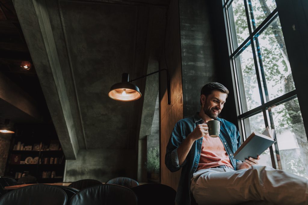 Portrait of cheerful breaded man drinking appetizing mug of liquid while reading interesting book opposite window indoor. Glad guy during leisure concept, digitalisierung, hotel trends